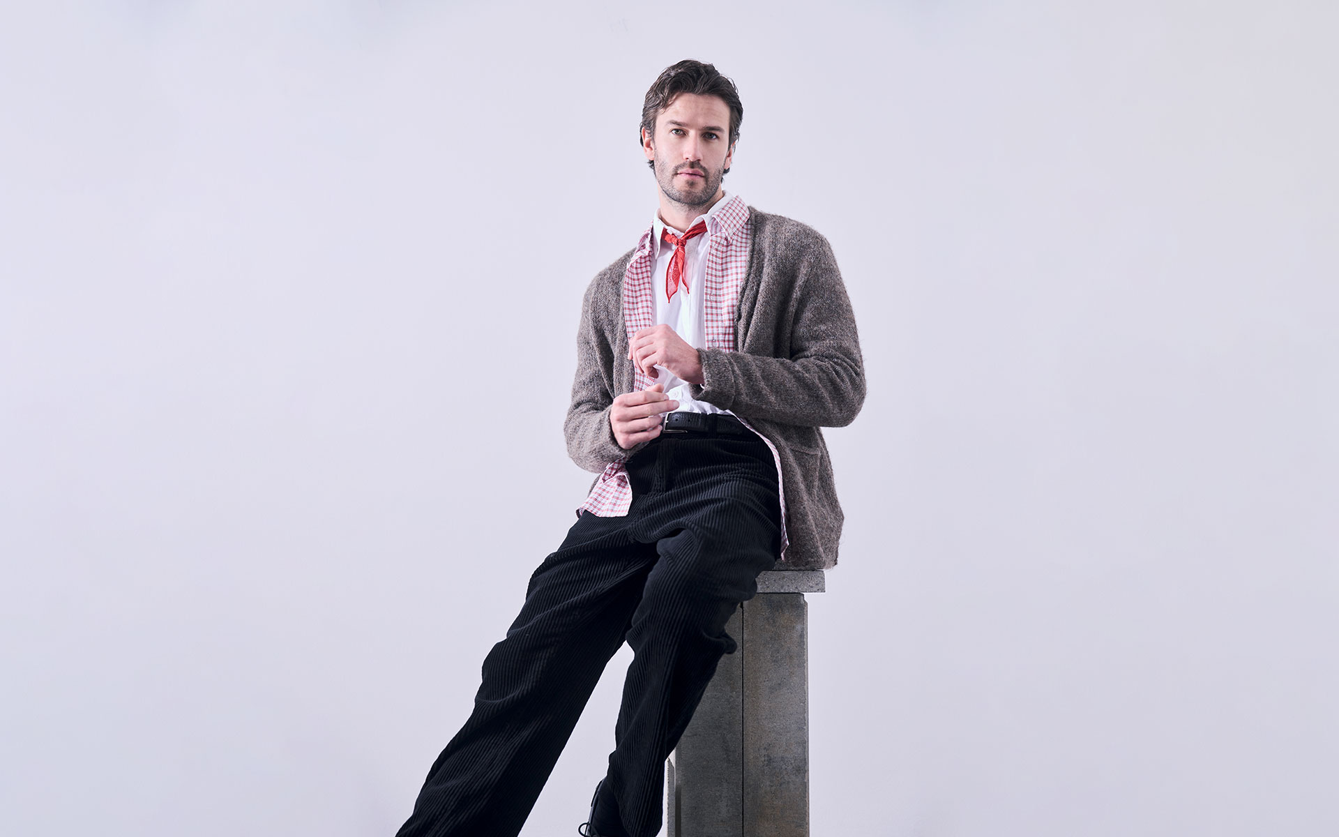 Ein Mann mit dunklem Haar und gestutztem Bart sitzt auf einem hohen, quadratischen Hocker vor einem schlichten Hintergrund. Er trägt eine braune Strickjacke, ein kariertes Hemd, eine schwarze Hose und ein rotes Halstuch und schaut selbstbewusst in die Ka