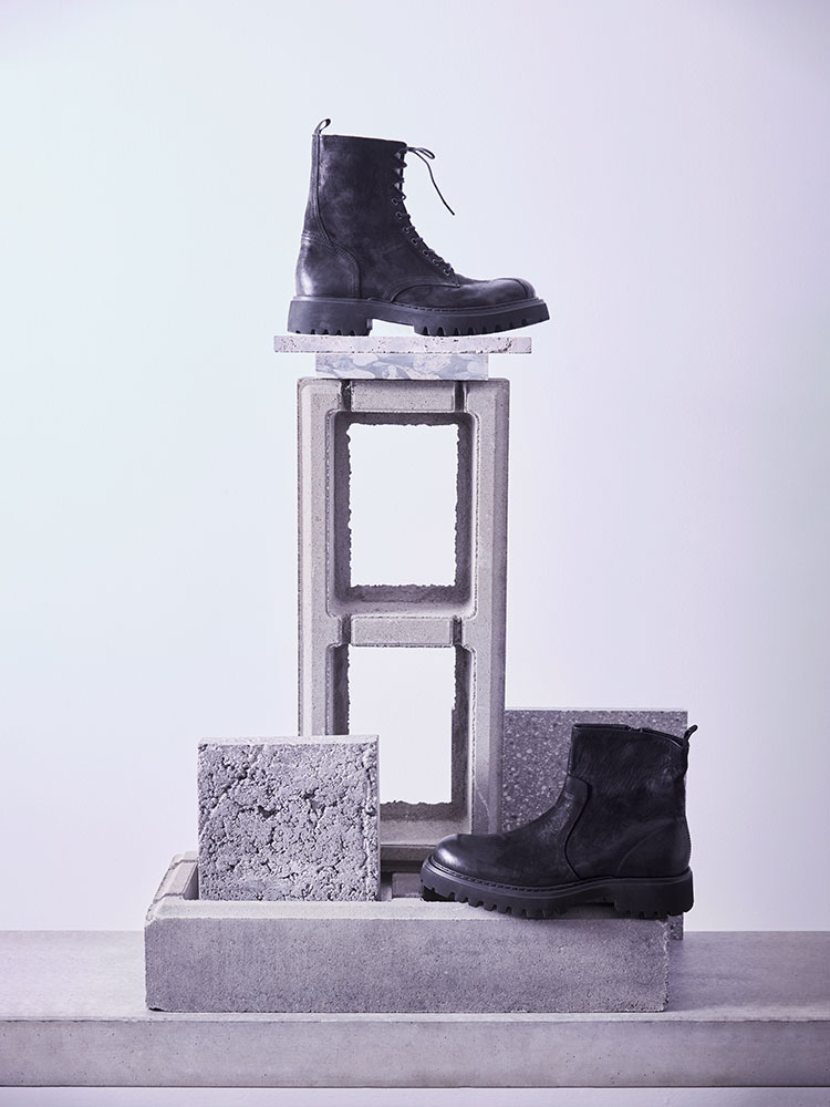 Zwei schwarze Schnürstiefel und Stiefel mit Reißverschluss, die auf und um gestapelte Betonblöcke und strukturierte graue Fliesen vor einem blass lavendelfarbenen Hintergrund ausgestellt sind.