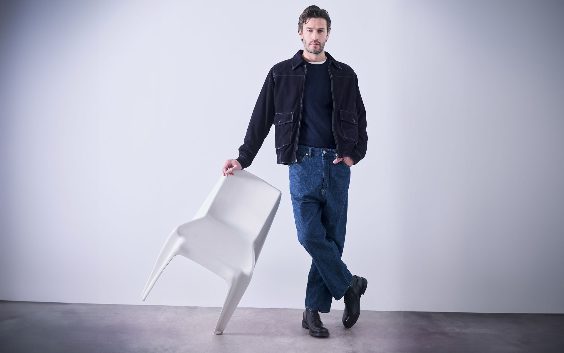 Ein Mann in schwarzer Jacke und blauer Jeans steht mit einer Hand in der Hosentasche und hält einen weißen, auf die Vorderbeine gekippten Stuhl vor einem schlichten, hellen Hintergrund.