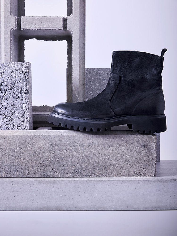 Eine schwarze Lederstiefelette mit klobiger Sohle wird auf einem Stapel von Betonblöcken und Schlackensteinen in einer minimalistischen Umgebung mit hellviolettem Hintergrund ausgestellt.