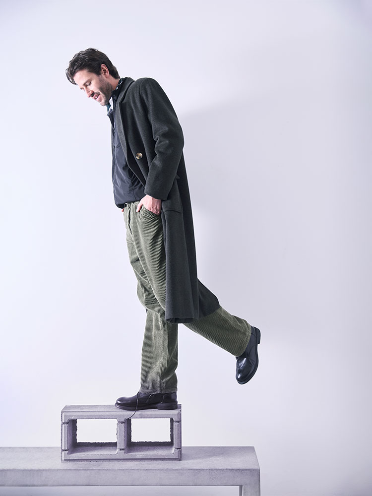 Ein Mann in einem dunklen langen Mantel, einem dunklen Hemd, einer grünen Cordhose und schwarzen Schuhen tritt von einem Stapel Betonblöcke auf eine weiße Plattform vor einem schlichten hellen Hintergrund.